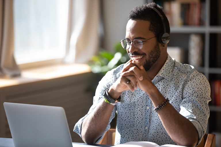 Educação Continuada - Feliz homem afro-americano milenar de óculos usando fones de ouvido, gostando de assistir a um webinar educacional no laptop.